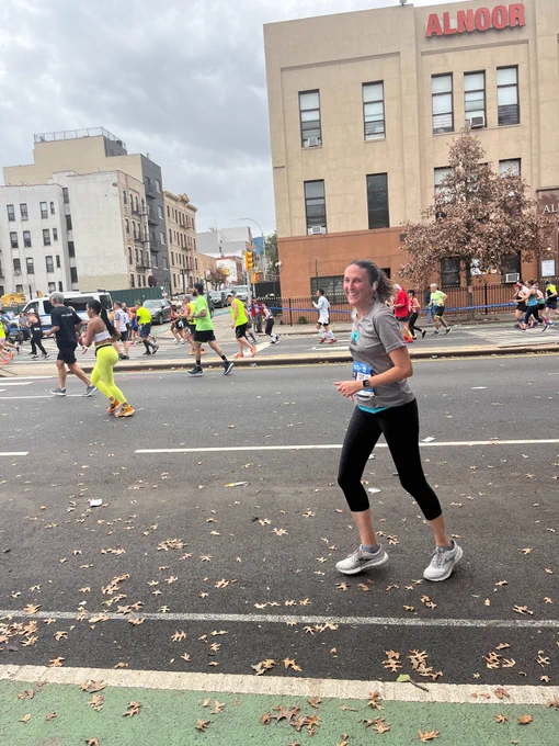 During the TCS NYC Marathon, a woman in black capri pants and a grey shirt with the PAWS NY logo turns to look at the camera, running in the street. Others run in the background as well, with buildings behind them as well.