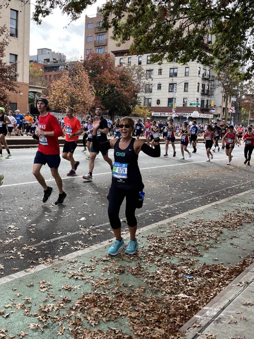 During the TCS NYC Marathon, a woman in black capri workout pants and a black tank top runs down the street, in the bike lane with leaves on the ground. She has both of her hands in the air and her expression is surprise at having just recognized a person in the crowd. Her name, Gigi, is on the front of her shirt in teal letters and she wears teal tennis shoes as well. Others run the race in the background.
