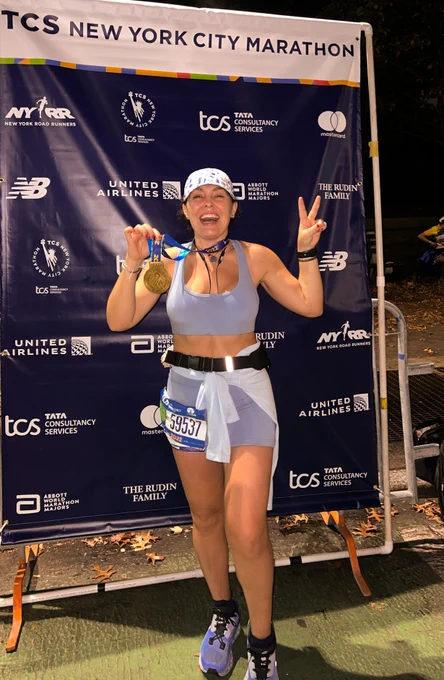 A happy young woman with an open-mouthed smile poses in front of a TCS New York City Marathon sign, holding her finisher's medal and giving a peace sign. She is wearing light grey shorts and a sports bra, with a white hat and black belt/pack. 