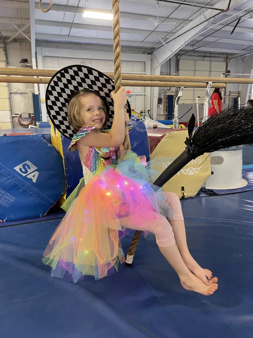 a participant in the Adaptive Gymnastics initiative in a gymnastics gym swinging on a rope wearing a witches hat and sitting on a broom