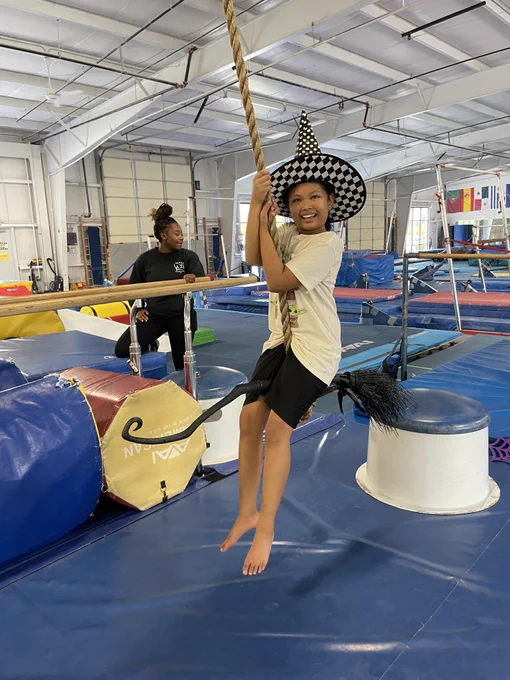 a participant in the Adaptive Gymnastics initiative in a gymnastics gym swinging on a rope wearing a witches hat and sitting on a broom