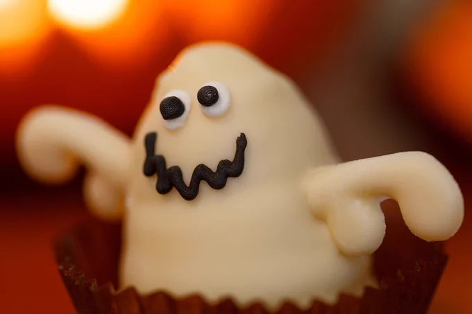 Photo of a ghost made of white chocolate in a dark brown paper cup. Its mouth and eyes are made of black icing. Background is diffused orange lighting.