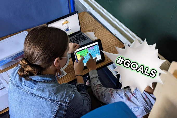 Picture of the back of an SLP and student sitting at a table. Both are pointing to an AAC device, which they are using to label emotions from a video playing on a laptop in front of them. A speech bubble that says #goals is covering the students head.