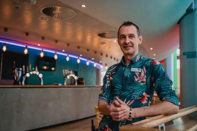 Alan Gibson, Customer Services Manager, stands with one arm leaning on a wooden rail in front of the bar area in ARC's No 60 café bar.