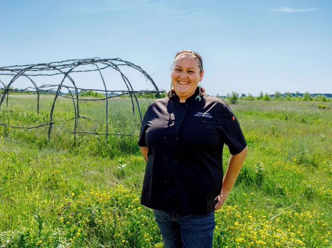 Elena Terry is standing in a bright green field in front of a hoop house, smiling and wearing a chef coat.