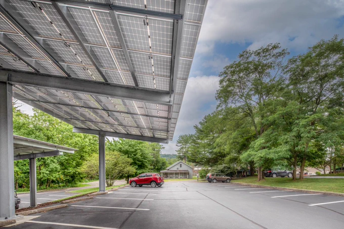 A solar carport provides shade and shelter to parked cars while generating electricity. The parking lot is flanked by green verdant trees.