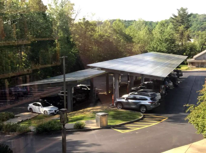 A photograph taken from above the solar carport shows the top of the solar array, the sun shining off its surface. Several cars are parked beneath benefiting from the shade.