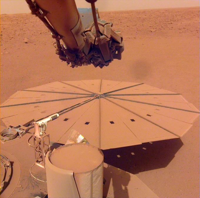 Photo from NASA’s Insight lander on Mars shows a portion of the lander and one of its solar panels covered in a thin layer of dust. Part of the lander’s robotic arm is visible at the top of the image. In the background, a dusty, rocky plain stretches to a flat horizon.