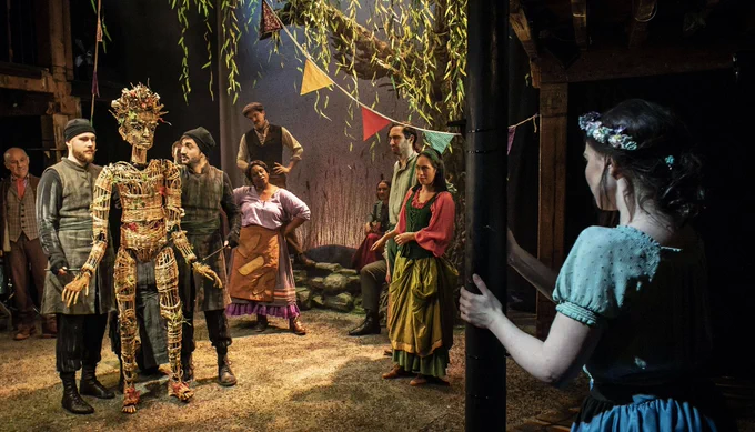 8 performers are on a gravel type stage. two holding a wicker  husband puppet. The other performances look at the wicker husband.