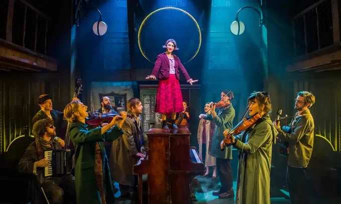 11 performers stand on a stage . One performer is wearing an outfit of  bright red skirt with a maroon jacket. Standing on top of two dark oak pianos. The other performances stand looking close by with a myriad of instruments