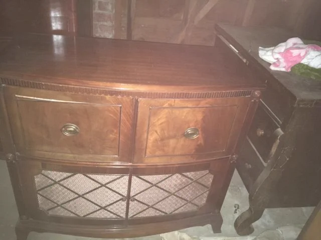 Photo of radio/player console. It’s a beautiful piece of polished hardwood furniture the size of a dresser, suitable for a living room or dining room. The two doors in the top half are for the radio (left) and record player (right). The bottom half has a fabric-covered screen where the speakers are.