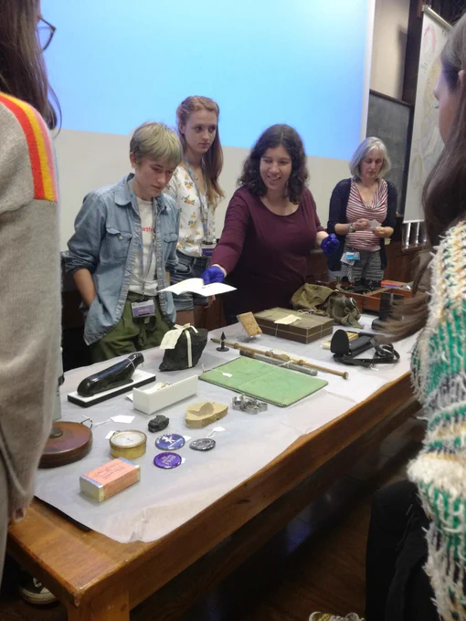 Table filled with intriguing objects. A group of teenagers look on while a museum staff member presents one of the objects.