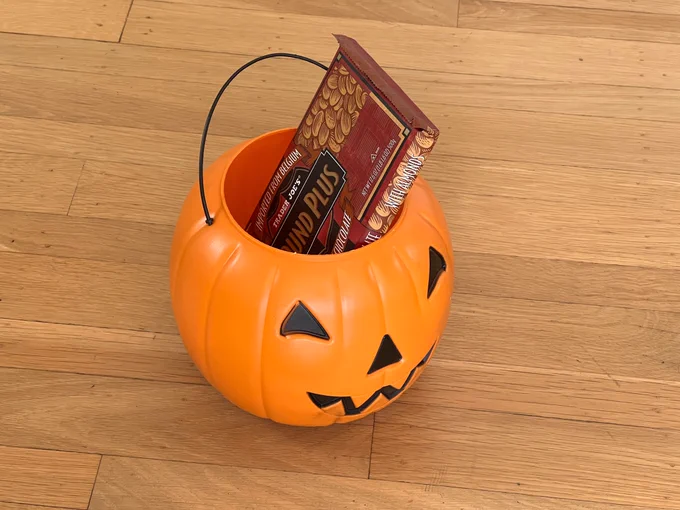 A standard-sized plastic jack-o-lantern trick-or-treating basket filled with candy, including a comically oversized 1 pound bittersweet chocolate bar sticking out of the top.