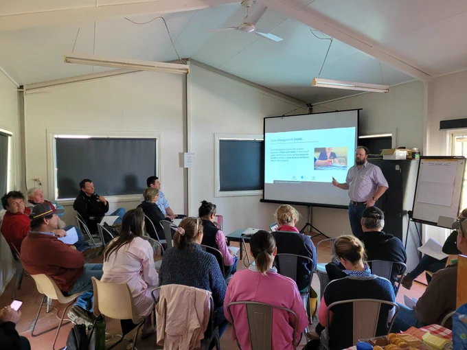 The photograph is taken at the Teelba community hall. It looks over the seated graziers to show a man talking through drought planning at the front of the room. November 2022