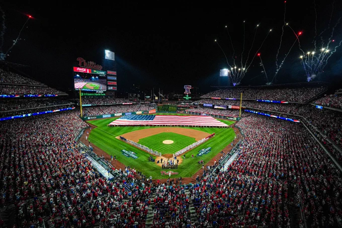 Photo of Citizens Bank Park filled with fans for Game 3 of the World Series. The American flag is in the middle of the field and the teams are lined up on both base lines. Fireworks are going off in the background.