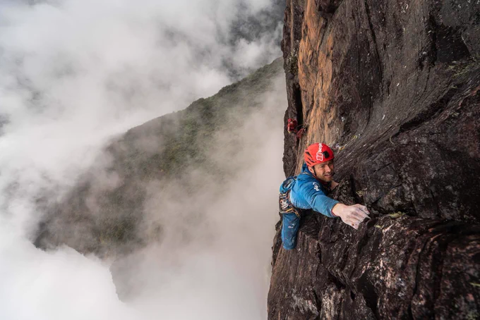 Climber Leo Houlding on the prow of Mount Roraima. Leo reaches for the next hold, behind him is a vertical drop into clouds and the Amazon rainforest.