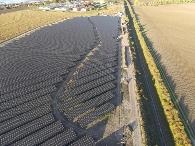 Das Foto zeigt eine Freiflächensolaranlage mit vielen blauen Photovoltaikmodulen. Im Hintergrund sind Gebäude des Dorfes Henschleben zu erkennen. Der Solarpark liegt zur Linken einer Bahntrasse.