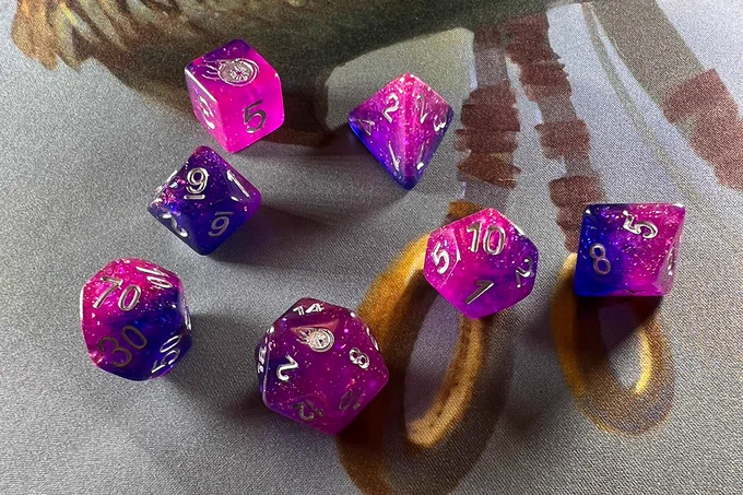 A set of 7 pink and purple polyhedral dice with silver numbers on a playmat. The high faces of two of the dice have a comet etched into them.