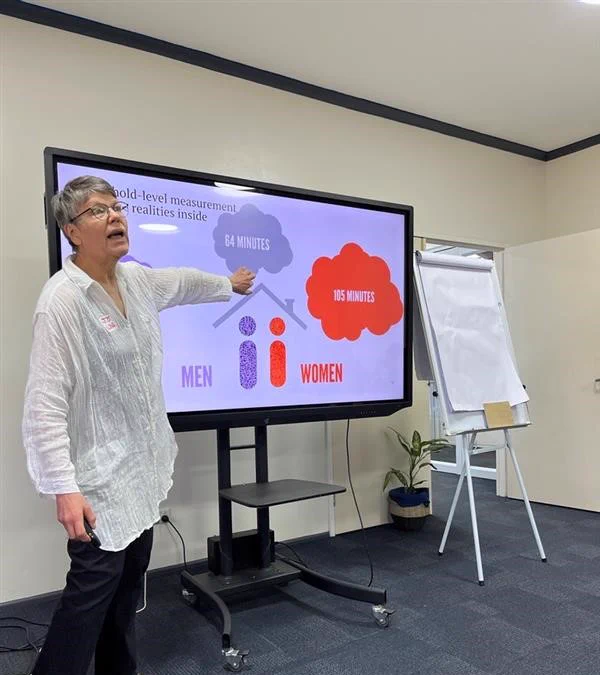 Jo is standing in front of a large screen presenting on gender data and poverty. She is white and has grey hair. She is wearing a long white top and black pants. She is pointing to the screen and is mid-sentence. 