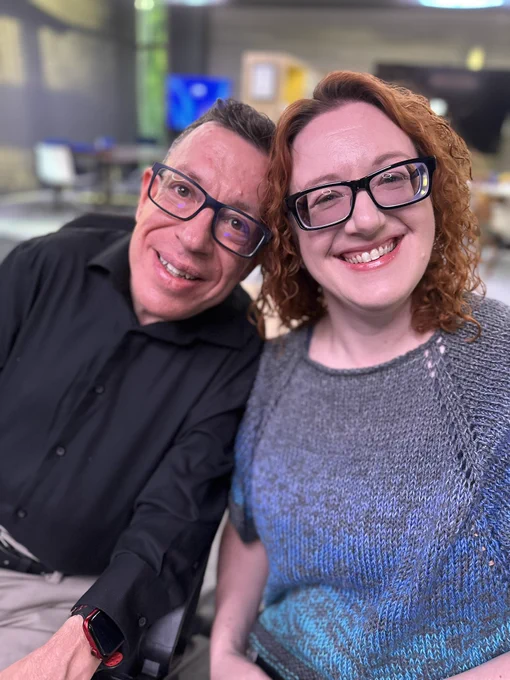 Two people sitting smiling at the camera. On right white female wearing glasses and strawberry blond hair, wearing blue green sweater. On left white male with glasses black shirt sitting in power chair