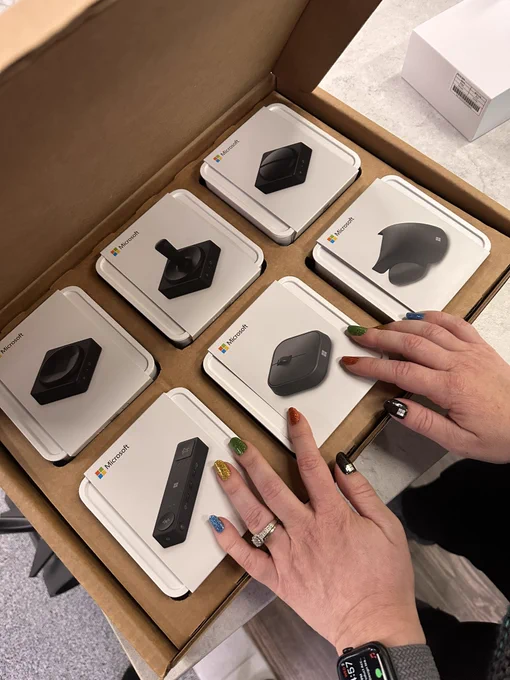 Cardboard box, opened containing 6 white boxes with different adaptive devices and accessories inside. The mouse, mouse tail, hub, button, joystick and pull button. Gabi’s hands are on 3 of the boxes with her fingernails painted in Microsoft colours - red green blue yellow and black thumbs with Microsoft logo