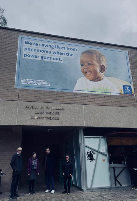 People standing under a University of Melbourne banner that reads "We're saving lives from pneumonia when the power goes out"