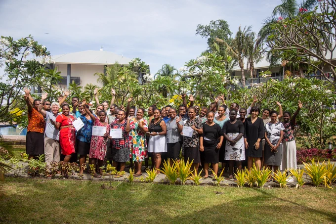 A large group of people holding their hands up in the air. Some people are holding certificates and everyone looks happy. They are outside and there is lots of grass and trees around. It looks tropical. 