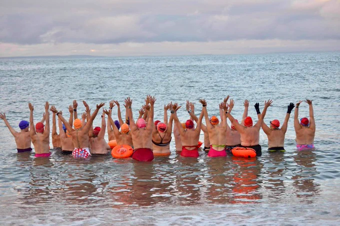 Colour photograph of 22 women in the sea. They stand with their backs to the camera with arms in the air and swimming costumes down to their waist. They symbolise the around 22 people diagnosed with lobular breast cancer in the UK everyday. 