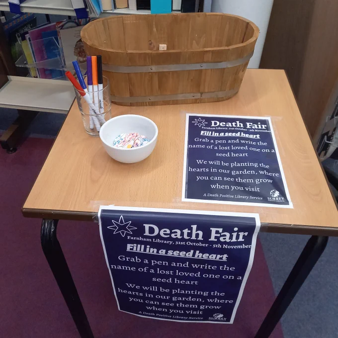 A photo of a table with seed hearts, pens, and a planter. A poster on the table reads:
Death Fair
Farnham Library, 31st October - 5th November 
Fill in a seed heart
Grab a pen and write the name of a lost loved one on a seed heart
We will be planting the hearts in our garden, where you can see them grow when you visit