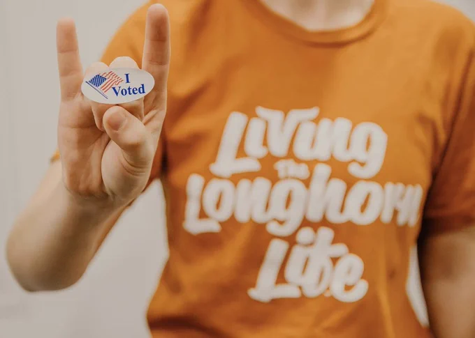 A student holds up an “I voted” sticker on their hand while making the “Hook ‘em, Horns sign.” The student’s t-shirt says “Living the Longhorn Life.”