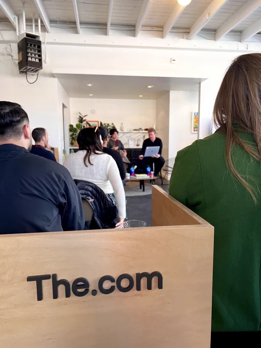 Caterina Fake talking with Clarke McKinnon, with the The.⁠com logo in the foreground