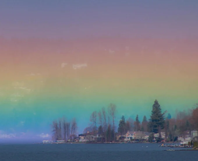 Photograph showing a lake shoreline with houses and trees to the right and flat blue water in the foreground, the sky above is in horizon bands of rainbow colour, green, yellow orange, red, pink and purple