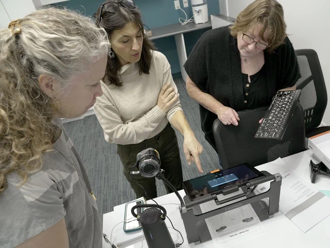 Three educators standing around the Prodigi Connect 12 by HumanWare,  with the PixPro camera attached to it.