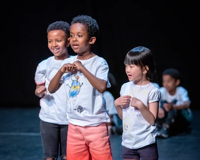 Three young people in white t-shirts. They are on a dark stage and are mid sentence