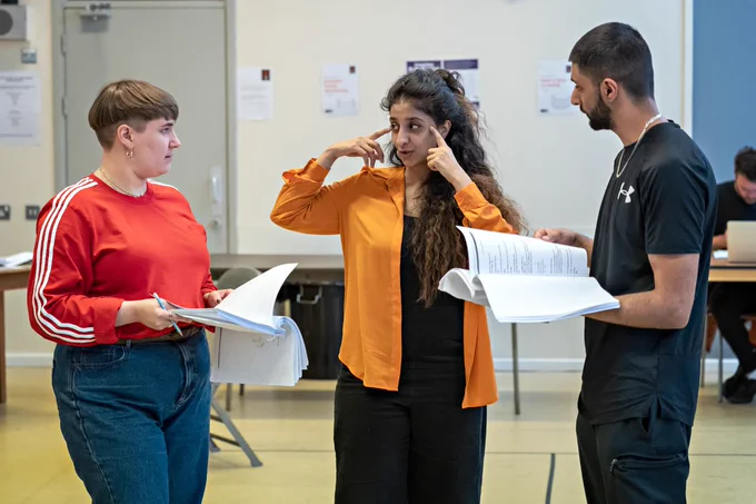 Three people in a rehearsal room. Two are holding scrips and one is pointing to their head