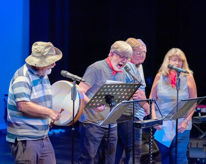 Four older people on a stage. They are singing and stood behind mic and music stands