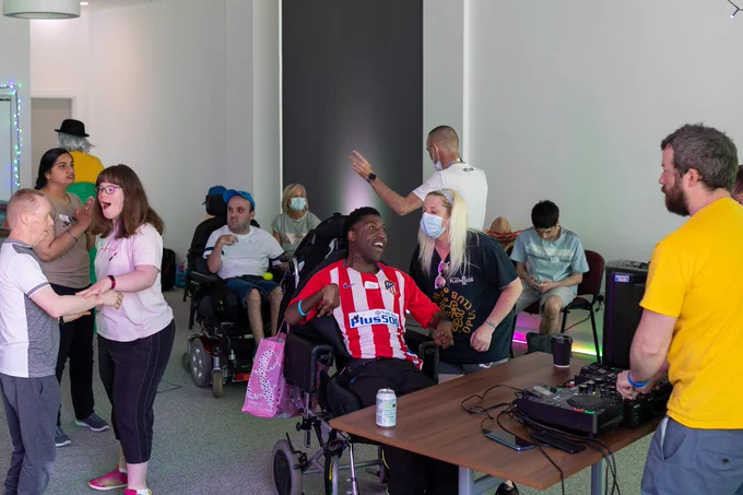 A room filled with people of all ages. They are dancing. A person in a yellow t-shirt stands behind a DJ stand. Two people are in wheelchairs
