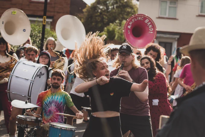 A young woman in a black tee-shirt dances a the front of a procession in the sun on a suburban street in Knowle West, Bristol, her hair flicking up in the air during the performance of SMOOSH! Behind her are rows of musicians including a clarinetist and a drummer playing a wheelchair-mounted kit.