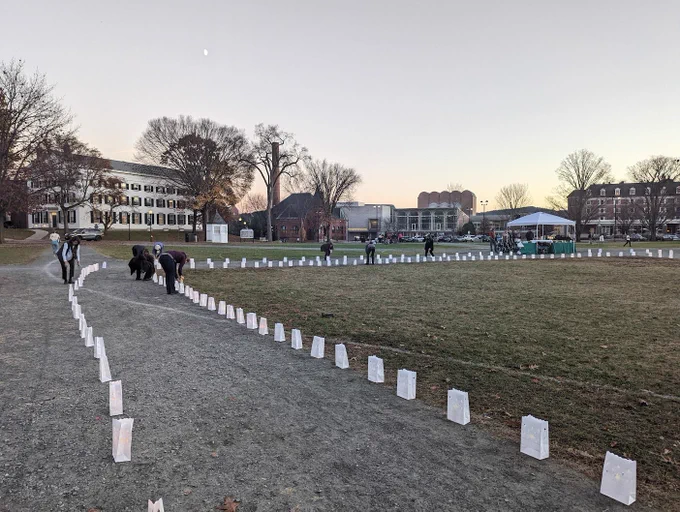 Volunteers light electronic candles in paper lanterns along a gravel walkway. The sun is setting but there is enough light to see by.
