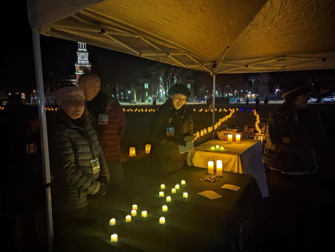 Members of the palliative care stand behind a greeting table, welcoming visitors. The table is covered with electric candles, and behind them stretches the lit pathway, glowing in the darkness. 