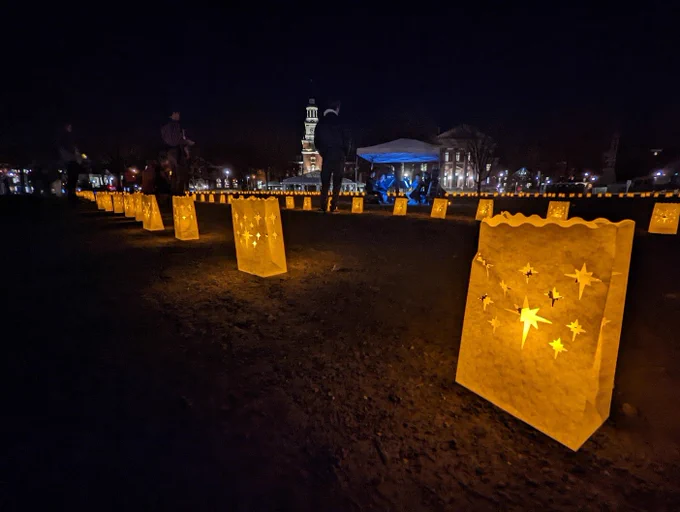 Luminaries glow with amber light along the pathway. A visitor stands on the path, listening to a volunteer string quartet in the center of the pathway. In the background, the Baker library tower is lit up. 