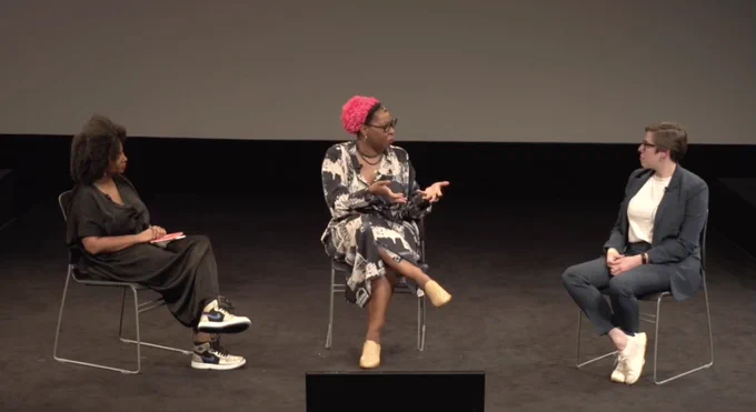 The three participants sit on chairs on a black stage. Theresa in the center faces Jane on the right as she asks her a question. A cue screen sits in the center front of the photo.