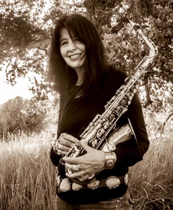 Joy Harjo smiling and holding a saxophone in a field. 