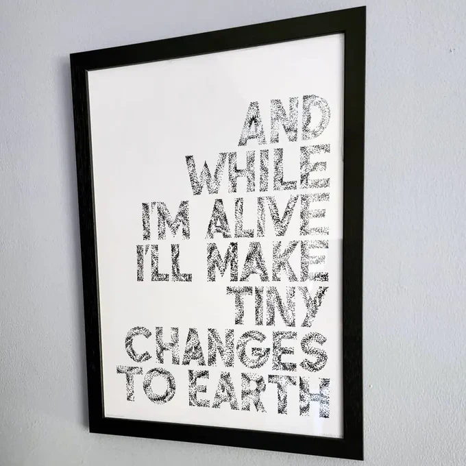 An art print of the line "and while I'm alive I'll make tiny changes to earth" from the Frightened Rabbit song "Head Rolls Off" in a black frame on a grey wall in Hanif's living room.