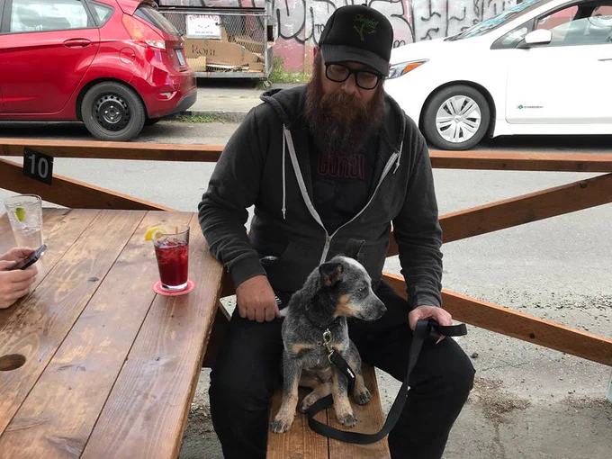 Aron Christensen, bearded and wearing a cap, is shown sitting at picnic table with his four month old puppy Buzzo on his lap