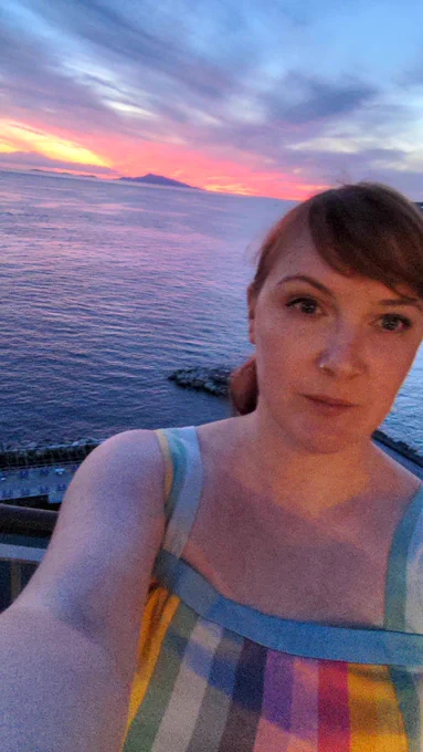 Me a white woman in her early 40s taking a photo of herself at the Sorrentine Bay with the sun setting over Capri in the background 