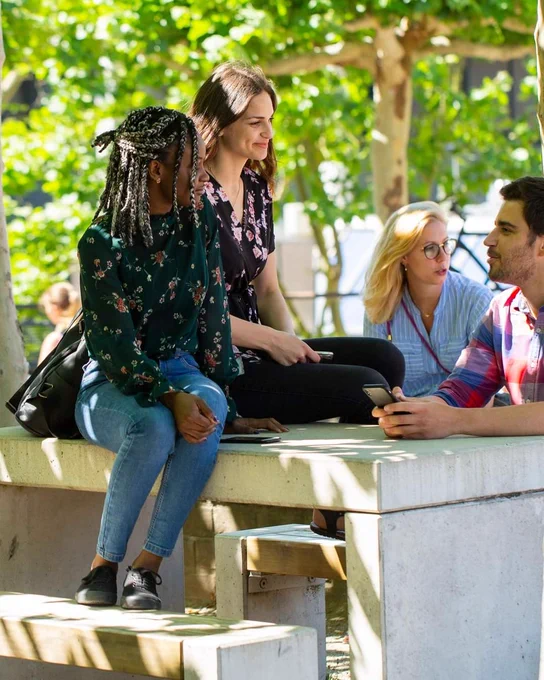 Studierende unterschiedlicher Hautfarben sitzen an und auf einem Tisch auf dem Campus und unterhalten sich. 