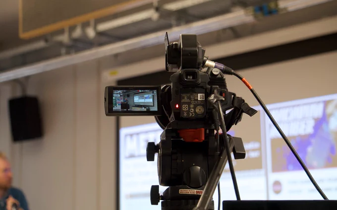 View through video camera with speaker and presentation screen in the background.