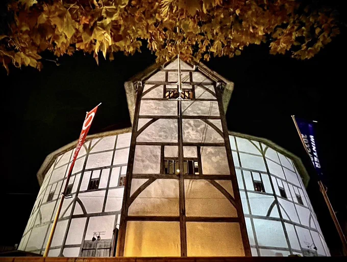 A nighttime view of the exterior of the Globe Theatre on Bankside. The camera looks sharply up at the tower of the Globe and it’s white lime plaster walls overhanging autumnal leaves from a tree reach into the top of the image.