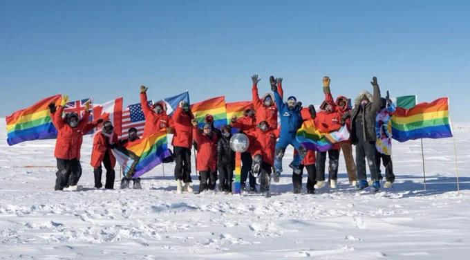 Sixteen people standing on ice and wearing cold weather gear cheer in front of several LGBTQ pride flags and national flags.
